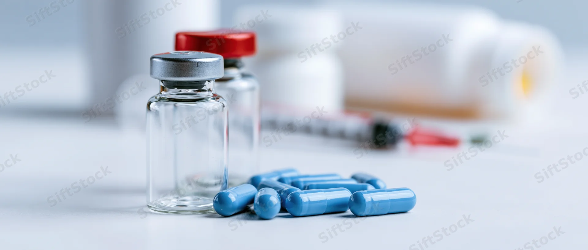 Medical vials and blue capsules arranged on a white surface with syringe in background