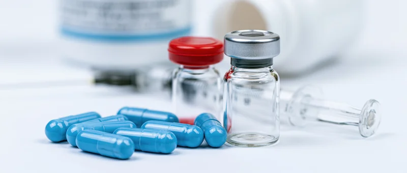 Blue capsules and glass vials with syringe arranged on white surface