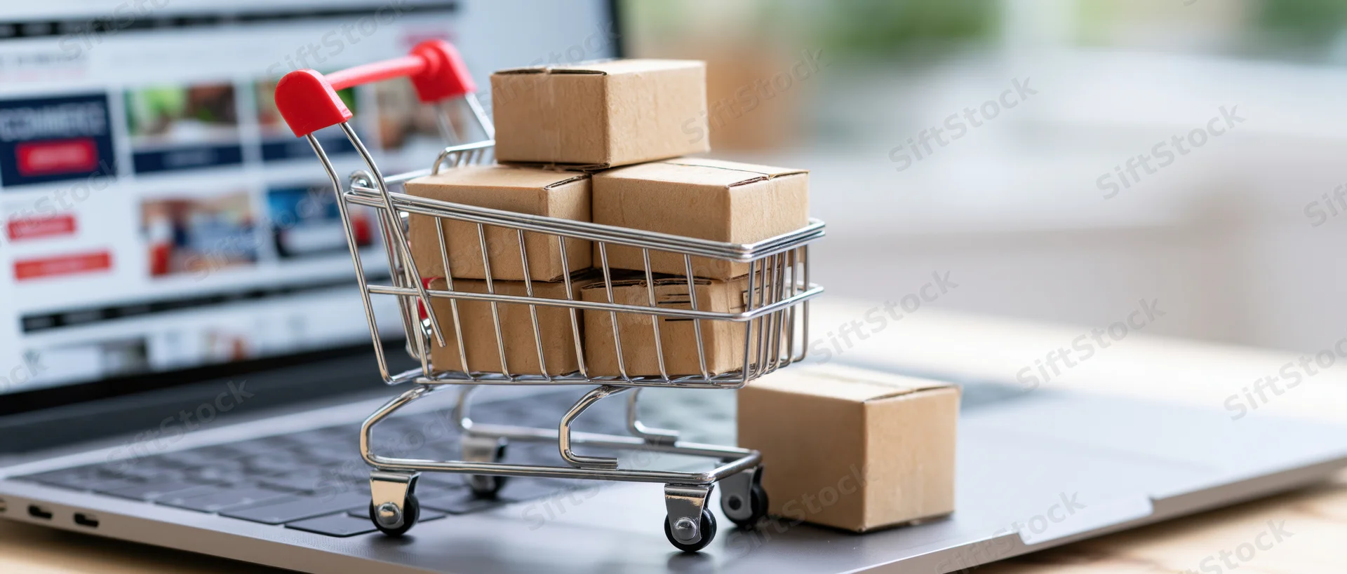 Miniature shopping cart filled with cardboard boxes on laptop keyboard near screen displaying online store