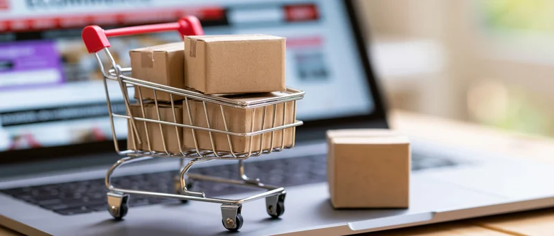 Miniature metal shopping cart filled with small cardboard boxes on laptop keyboard with online store screen in background