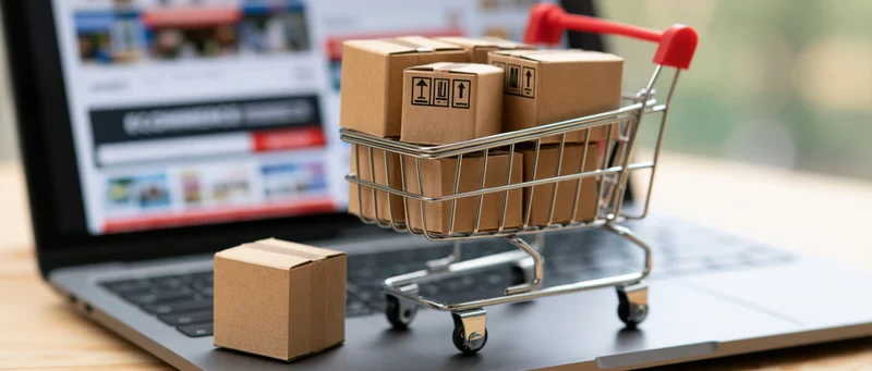 Miniature shopping cart filled with cardboard boxes on laptop keyboard with e-commerce website in background
