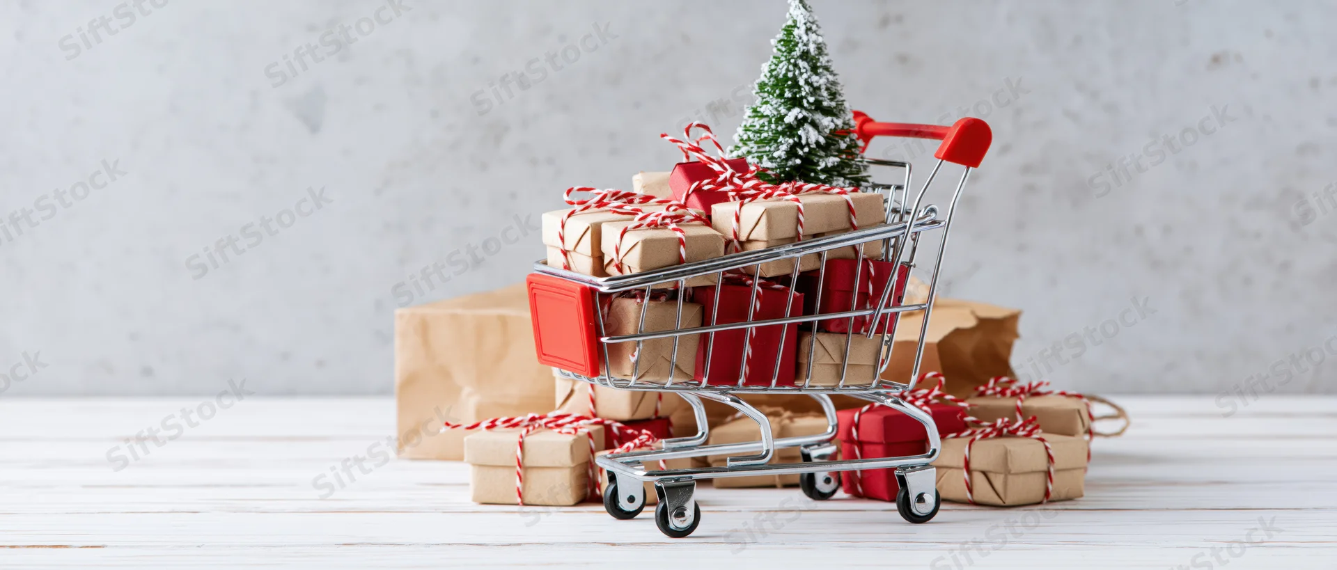 Miniature shopping cart filled with wrapped gifts and a small christmas tree on a white wooden surface