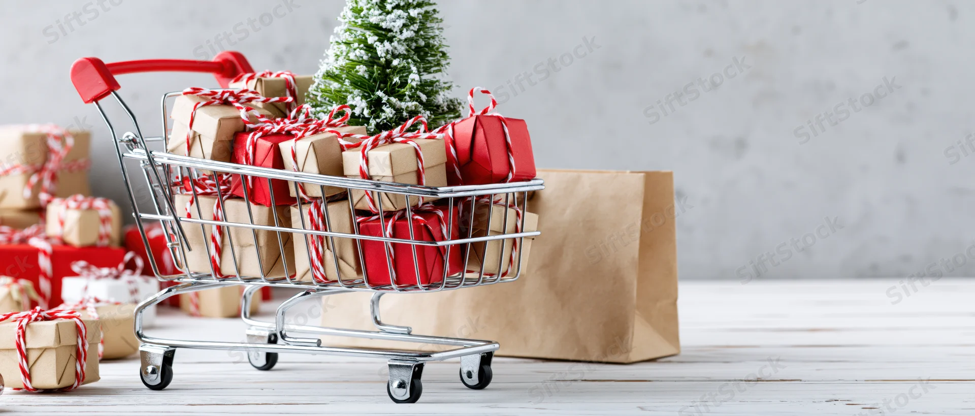Miniature shopping cart filled with wrapped gifts and a small christmas tree on a wooden surface beside a paper bag