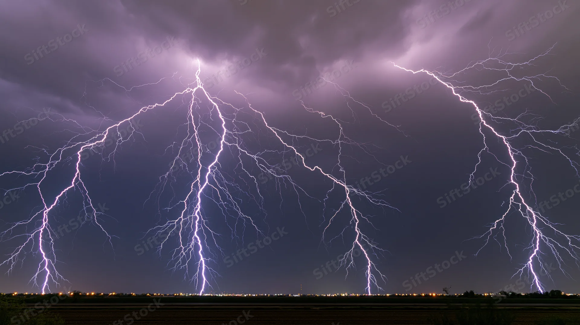 Multiple lightning bolts striking across a dark stormy sky over a rural landscape with distant city lights at night