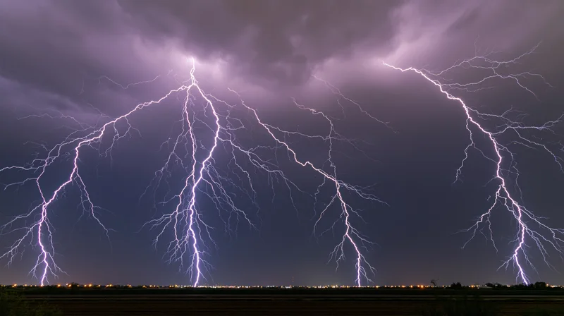 Multiple lightning bolts striking across a dark stormy sky over a rural landscape with distant city lights at night