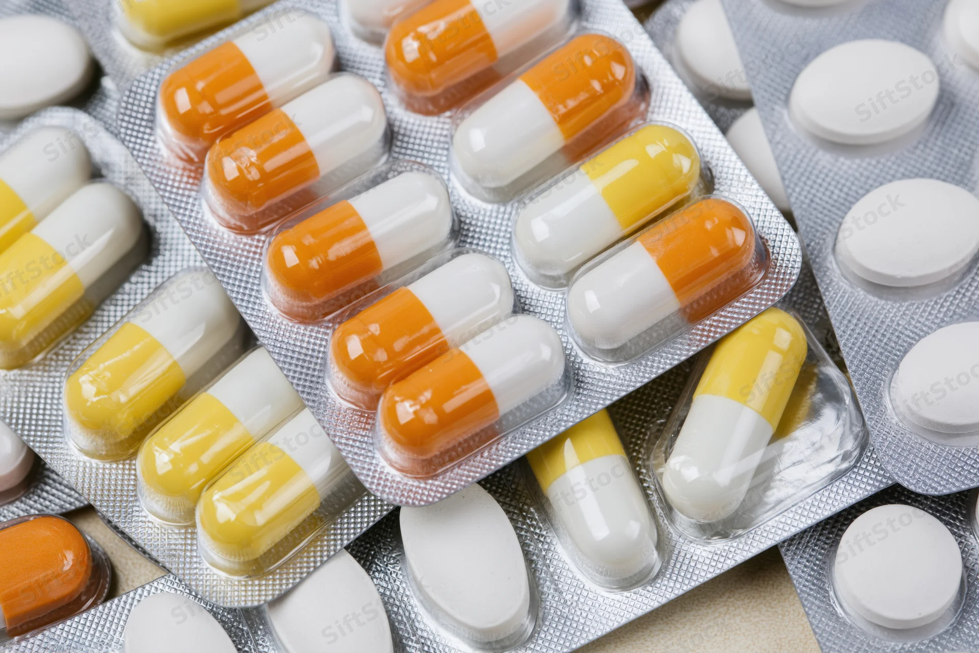 Multiple pharmaceutical blister packs filled with orange-white and yellow-white capsules arranged in overlapping layers