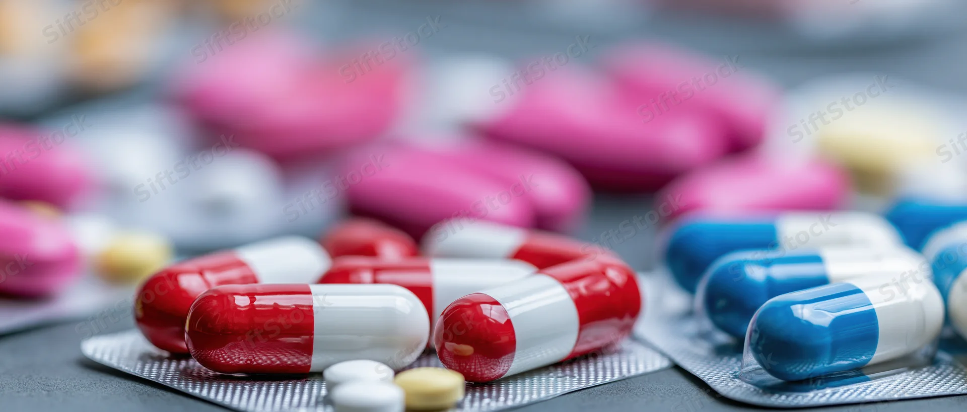 Red and white capsules with blue and pink tablets scattered on a gray surface in a blister pack arrangement