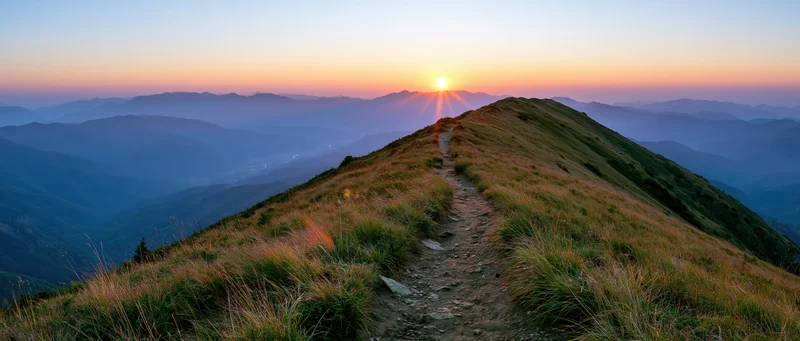 Narrow dirt trail winding along mountain ridge toward setting sun at dusk