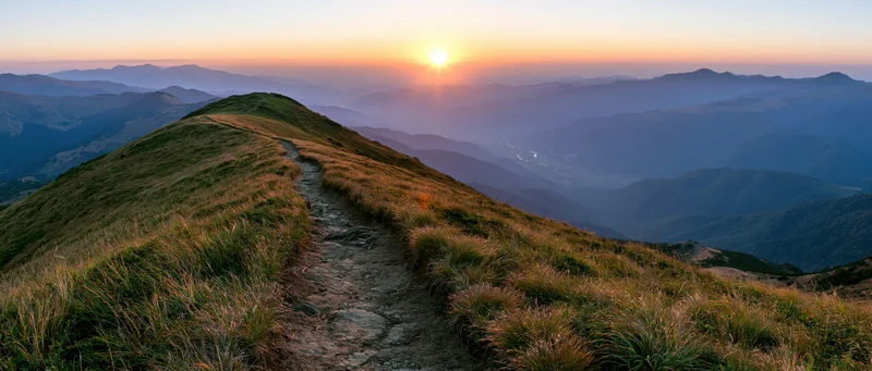 Narrow dirt trail winding along a mountain ridge toward a low sun at sunset