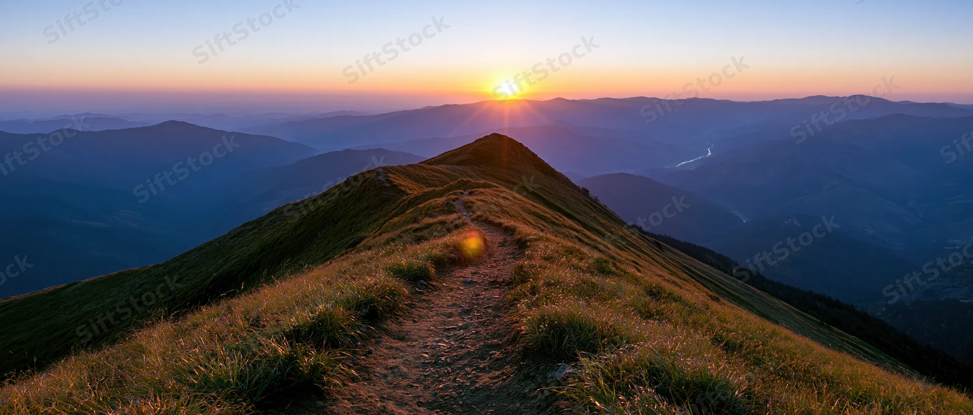 Narrow dirt trail winding along a mountain ridge toward a low sun at sunset