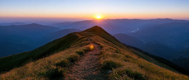 Narrow dirt trail winding along a mountain ridge toward a low sun at sunset