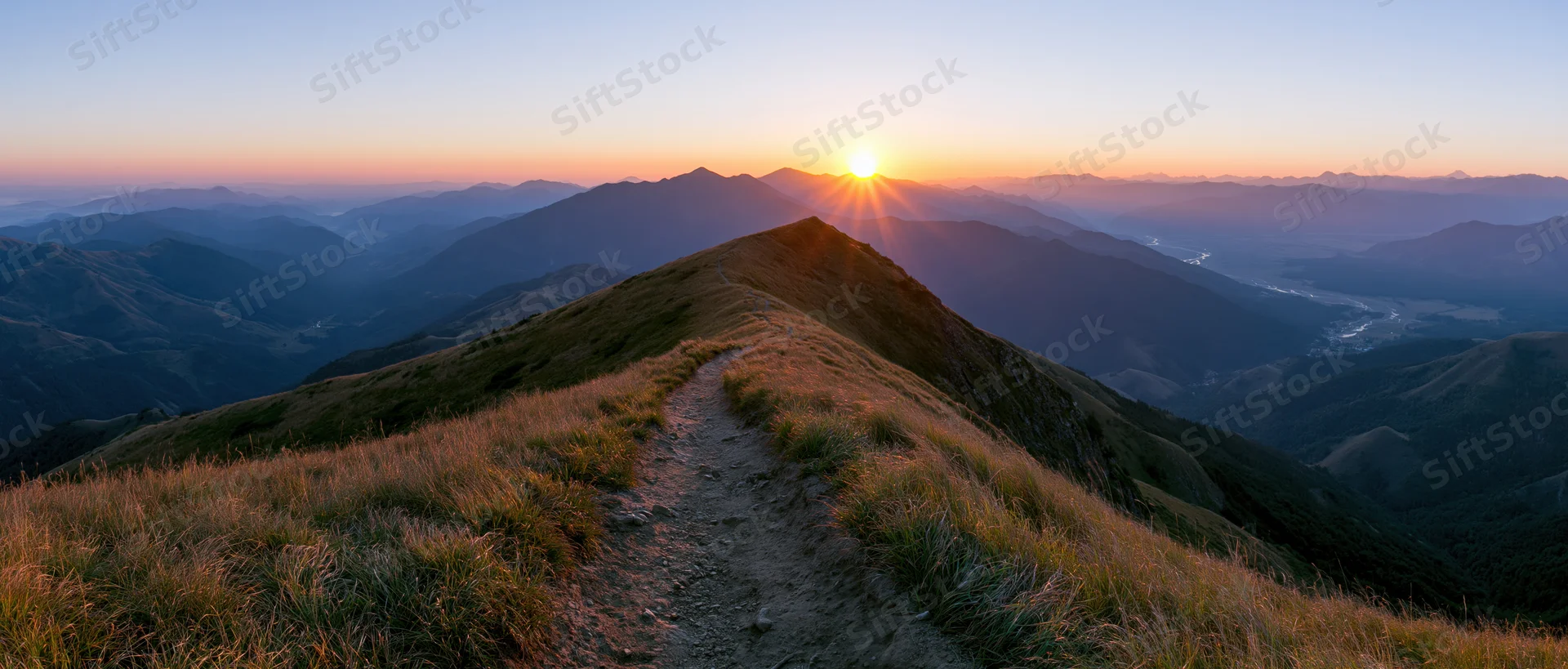 Narrow dirt trail winding along a mountain ridge toward a low sun at sunset