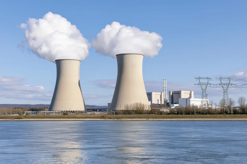 Two large concrete cooling towers emitting steam at a nuclear power plant beside a river under a clear blue sky
