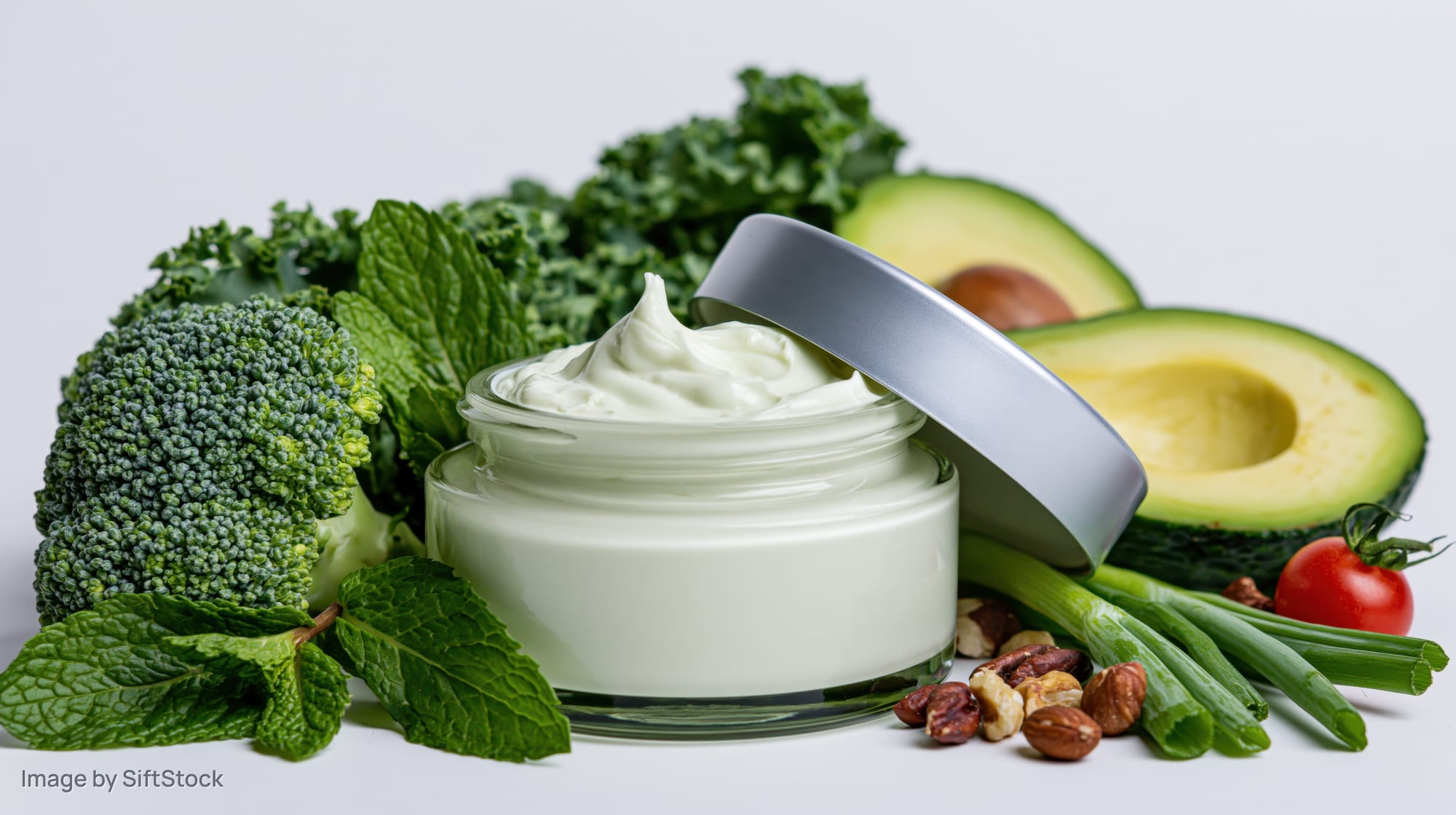 Open glass jar of white face cream with silver lid surrounded by fresh vegetables and herbs on white background
