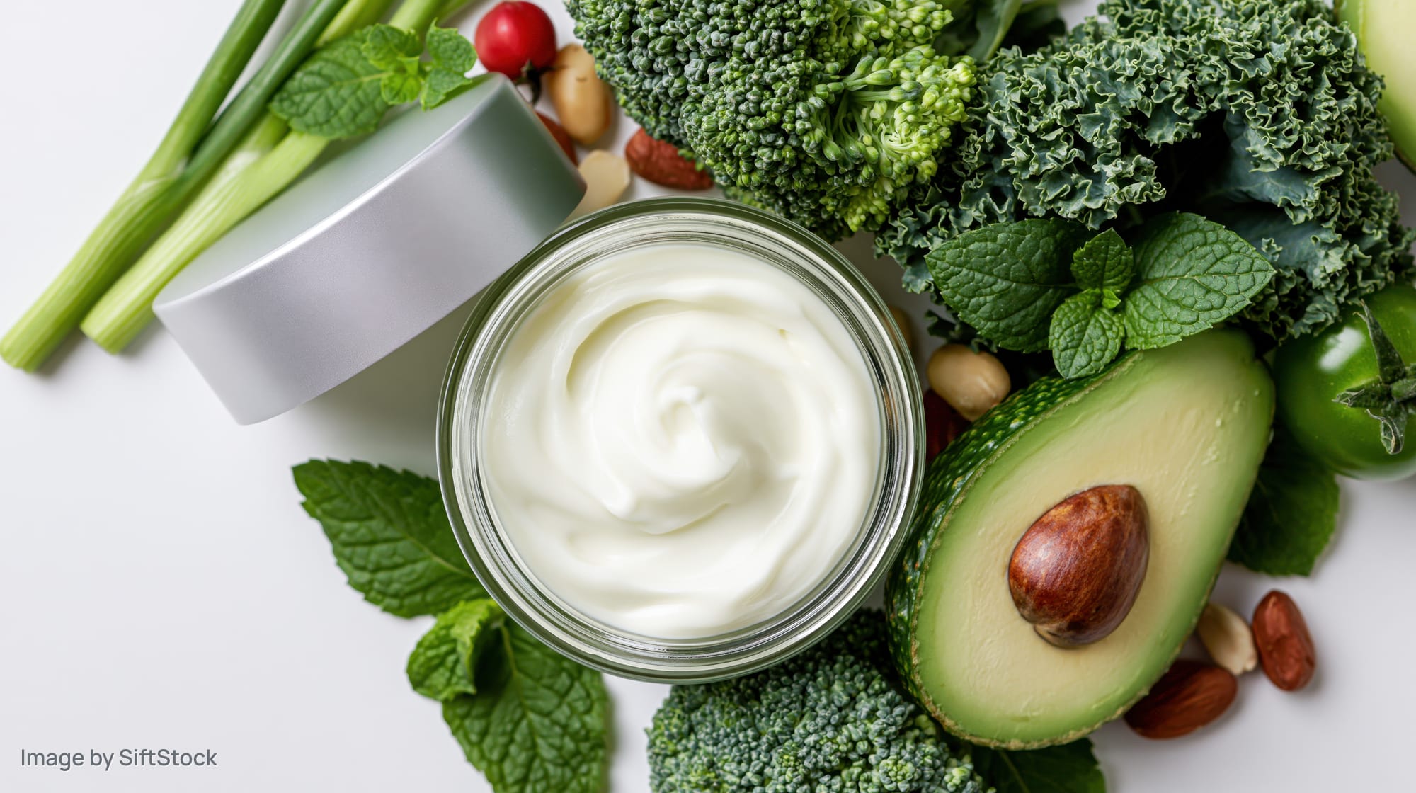 Open glass jar of white face cream surrounded by fresh vegetables and herbs on white surface