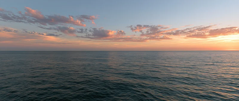 Open ocean at sunset with calm water reflecting warm orange sky and scattered clouds along the horizon