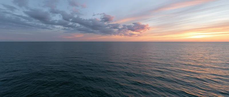 Open ocean at sunset with calm water reflecting warm orange sky and scattered clouds along the horizon