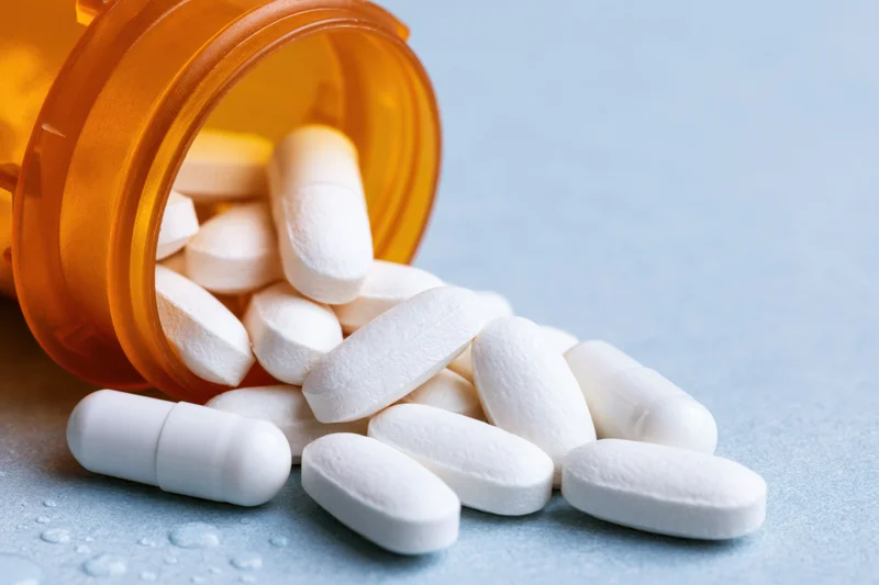 White pills and capsules spilling from an orange prescription bottle onto a blue surface