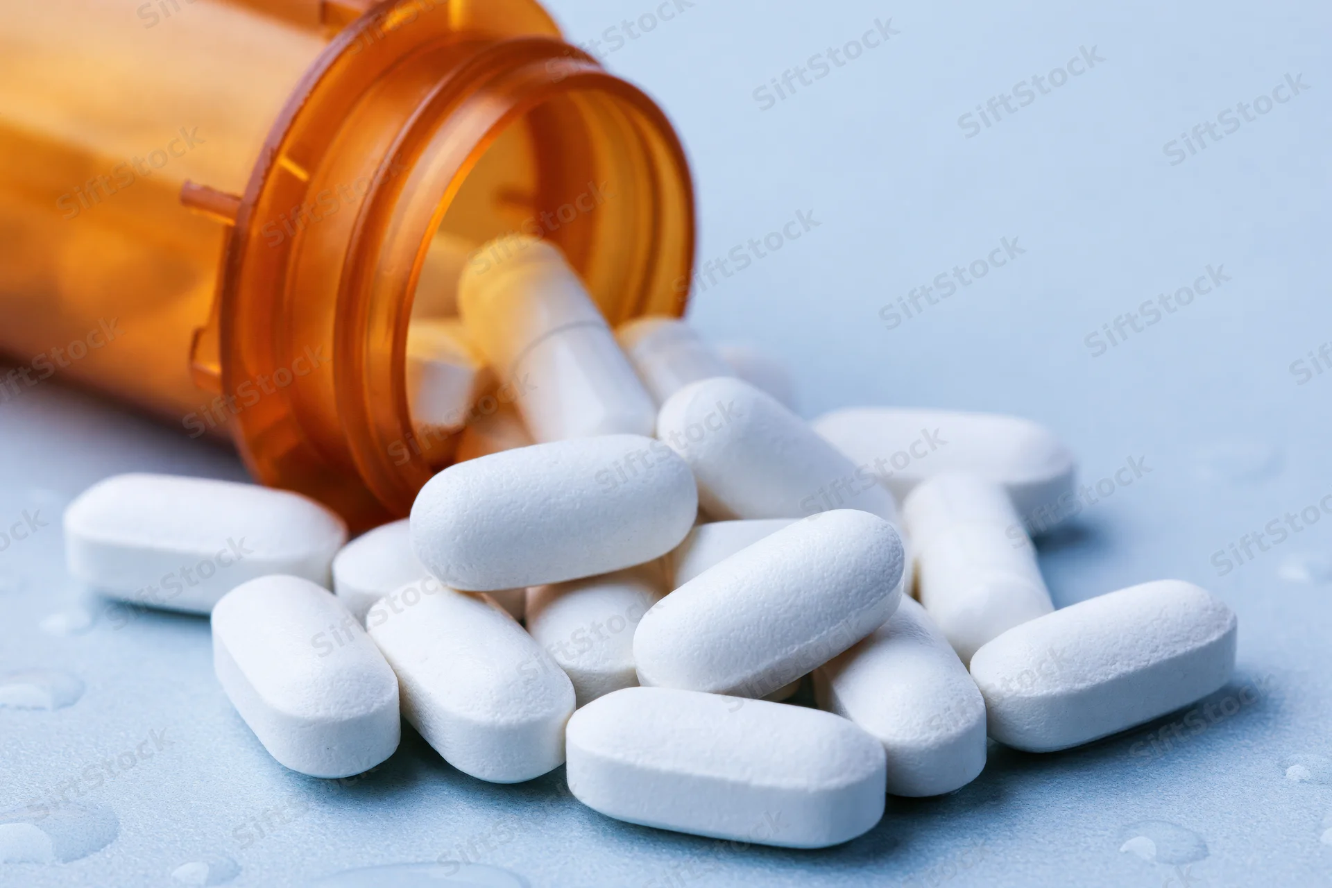 White oval pills spilling from an orange prescription bottle onto a light blue surface