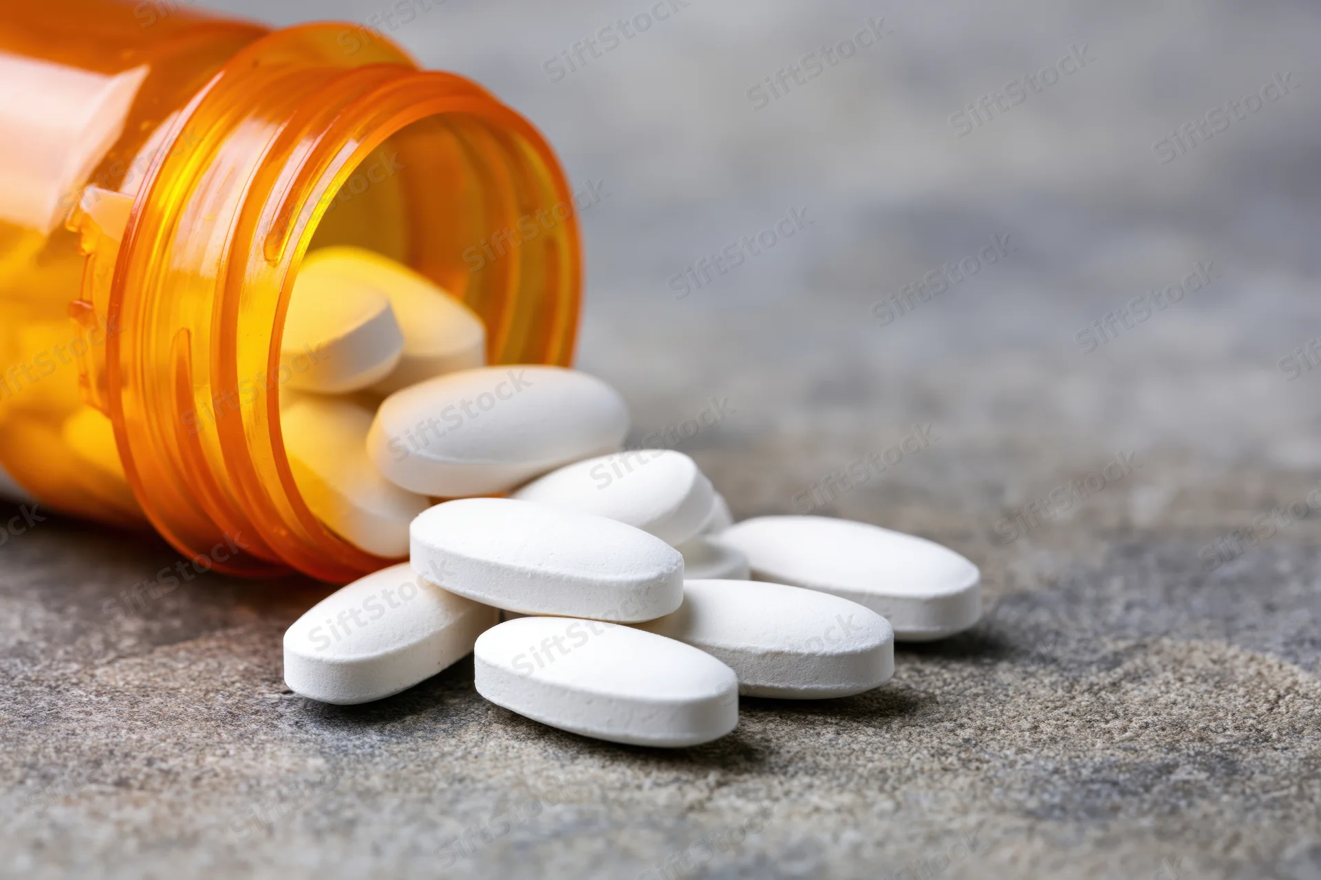 White oval pills spilled from an orange translucent prescription bottle onto a gray stone surface