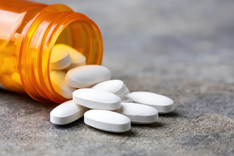 White oval pills spilled from an orange translucent prescription bottle onto a gray stone surface