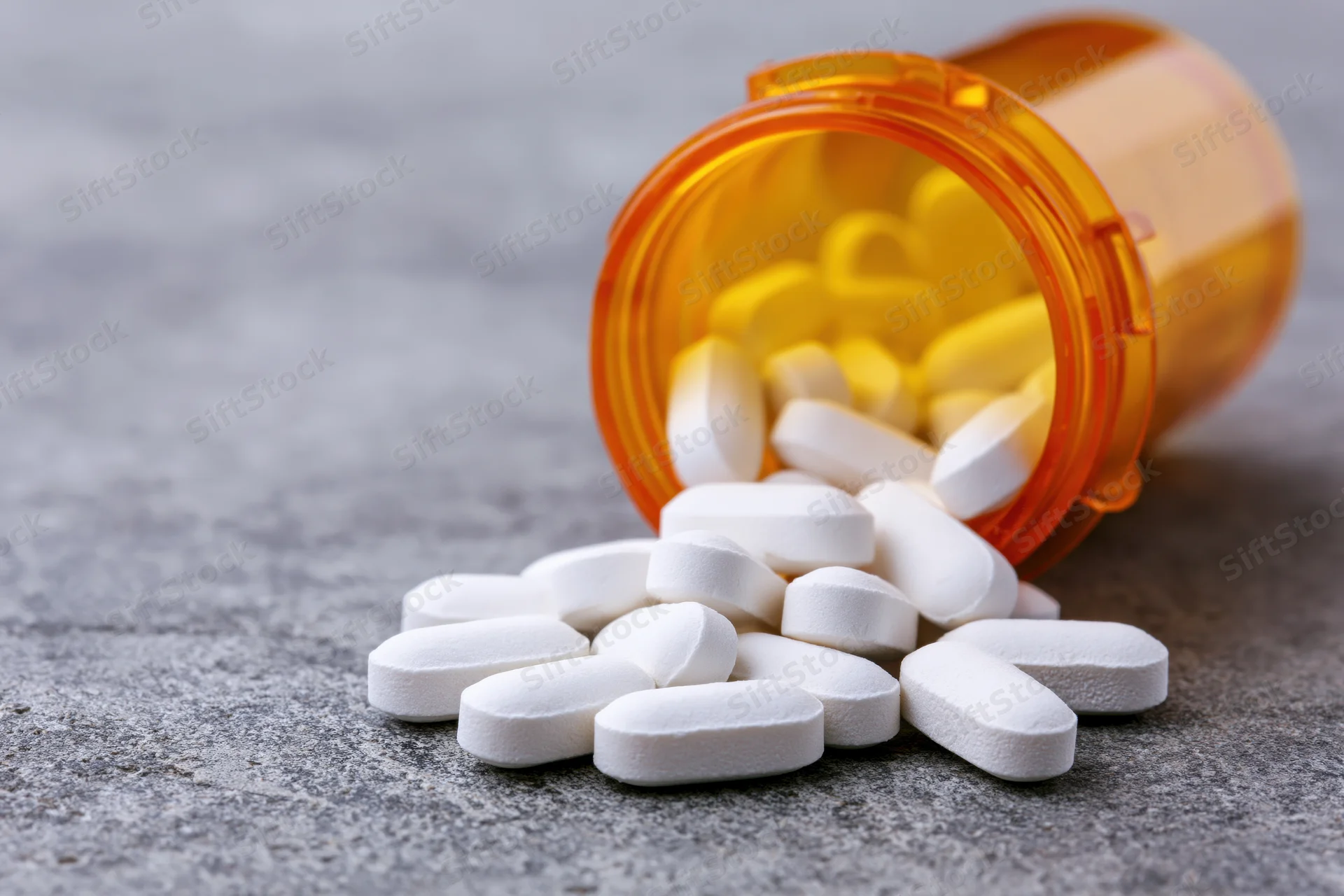 White oval pills spilled from an orange translucent prescription bottle on a gray surface