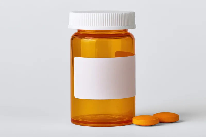 Orange translucent prescription pill bottle with blank label and two orange tablets beside it on white background