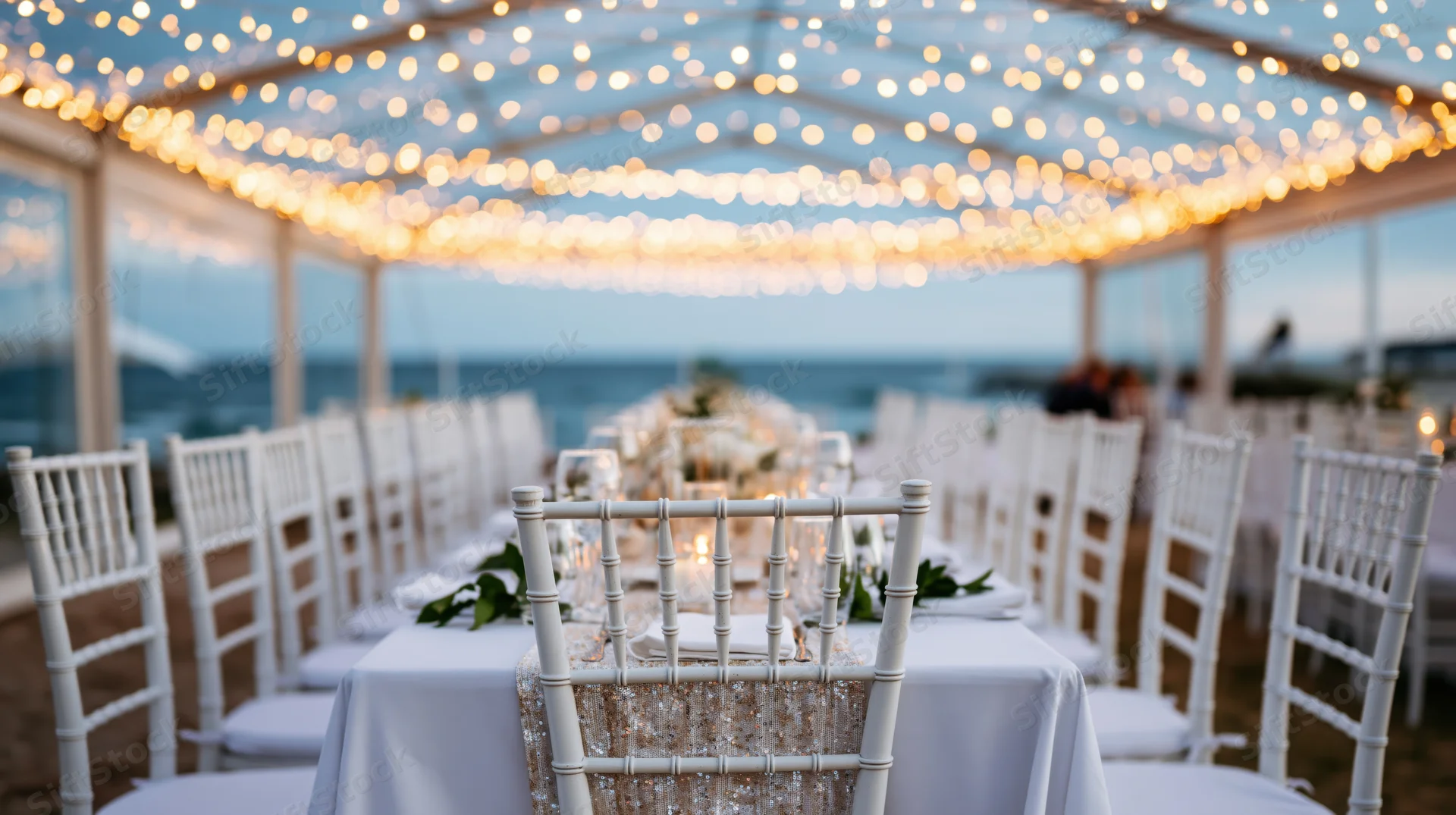 Wedding reception table set under clear tent with string lights overlooking ocean at twilight