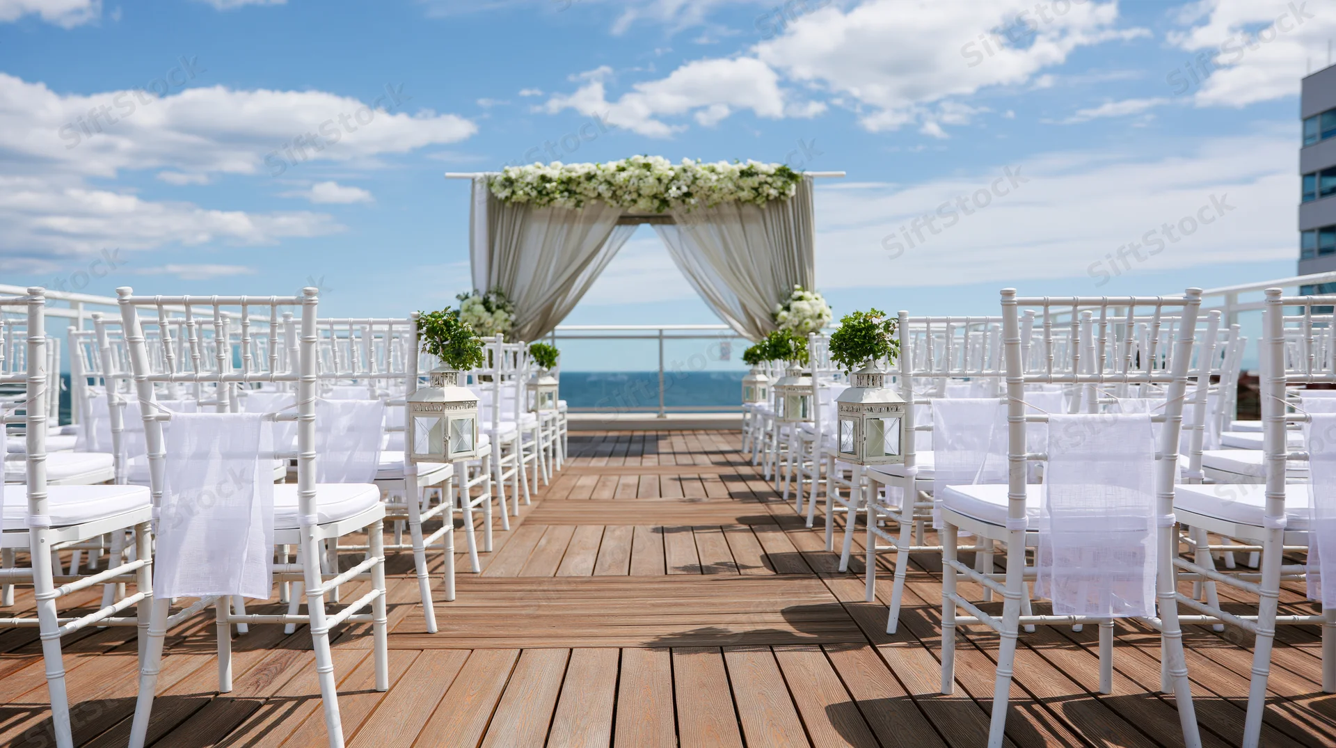 White chiavari chairs arranged on wooden deck facing floral arch with ocean view under blue sky