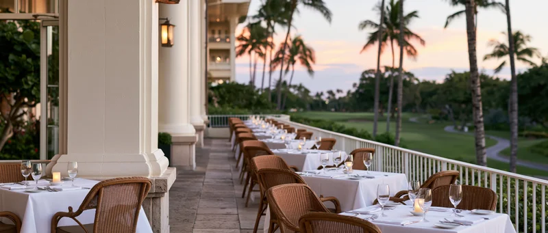 Empty dining tables set on a covered terrace overlooking a tropical golf course at sunset