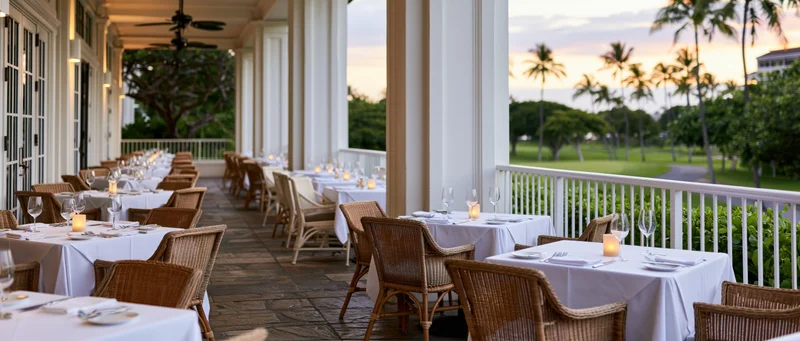 Empty dining tables set with candles and glassware on a covered terrace overlooking a tropical garden at sunset