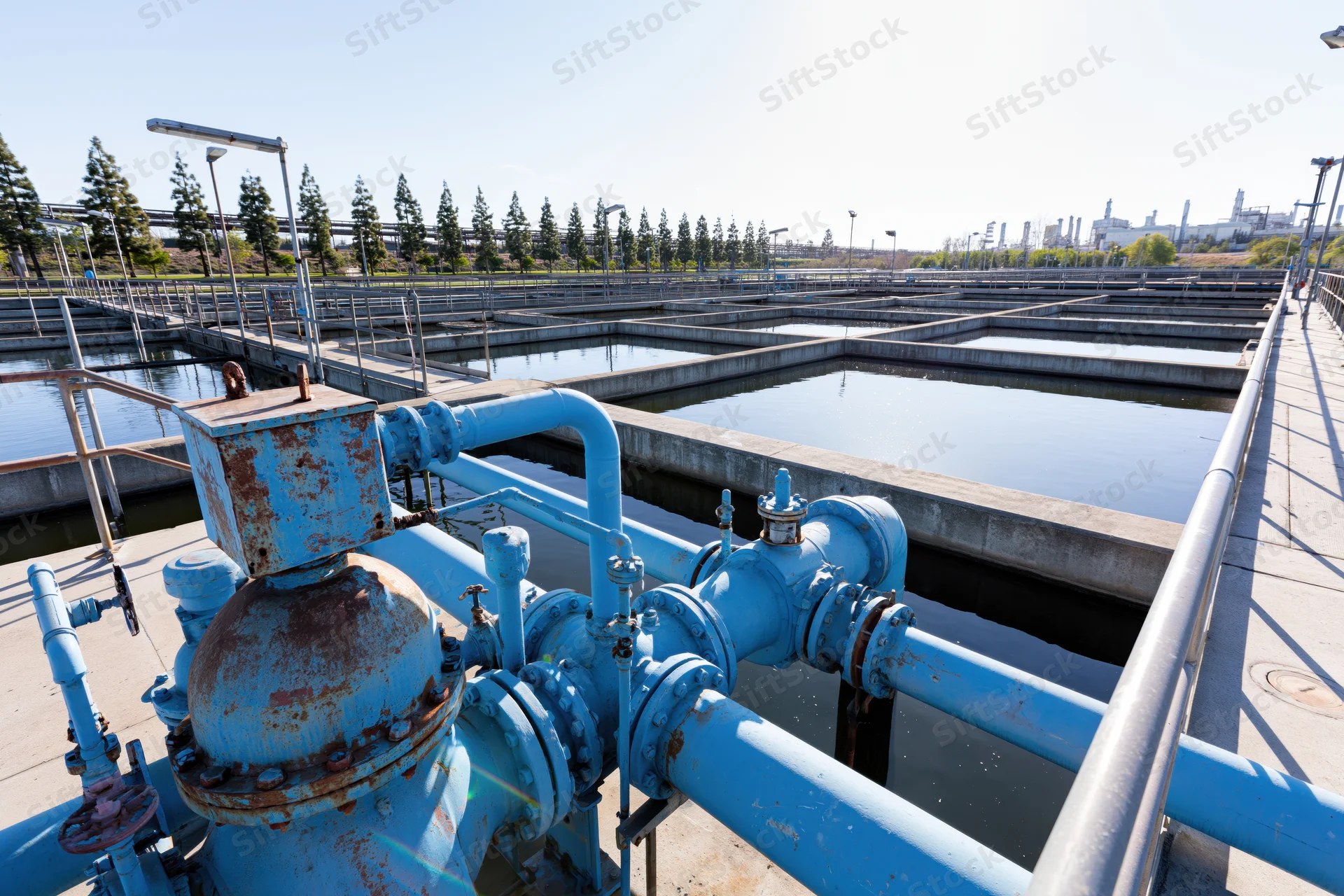 Blue pipes and valves at a wastewater treatment plant with concrete settling tanks under a clear sky