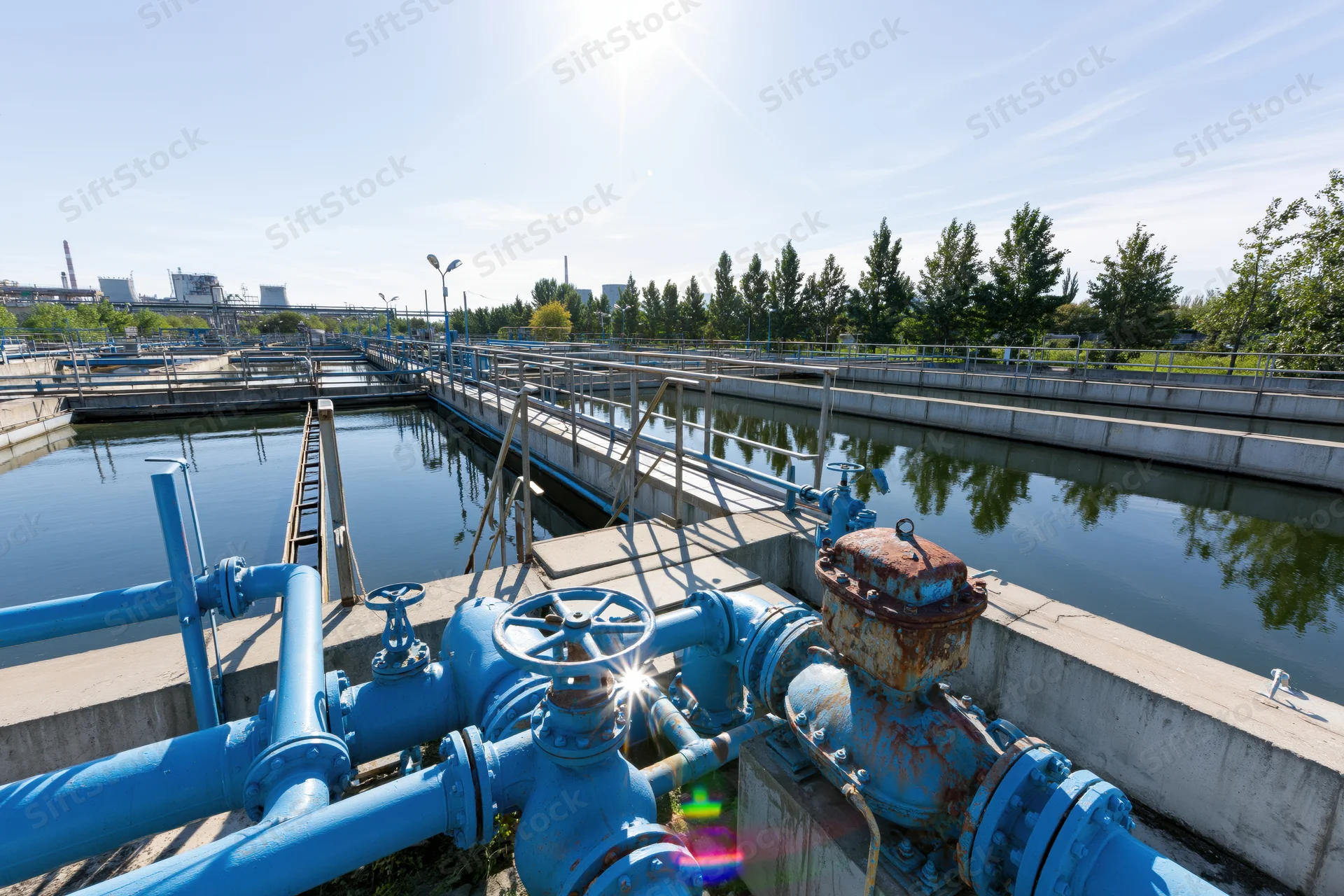 Wastewater treatment plant with blue pipes and settling tanks under bright sunlight