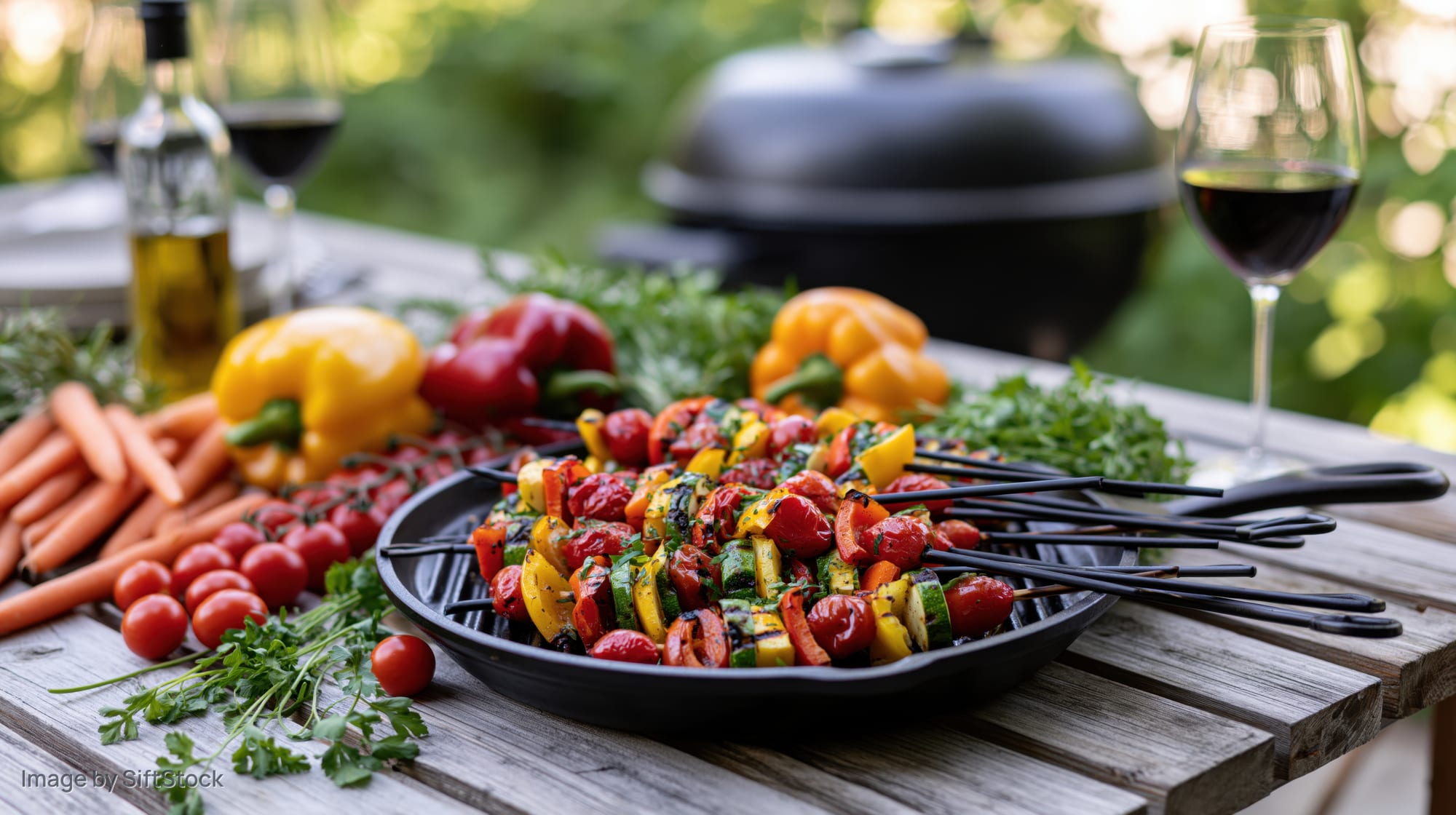 Vegetable skewers arranged on a black grill pan on a wooden patio table with wine and fresh produce around