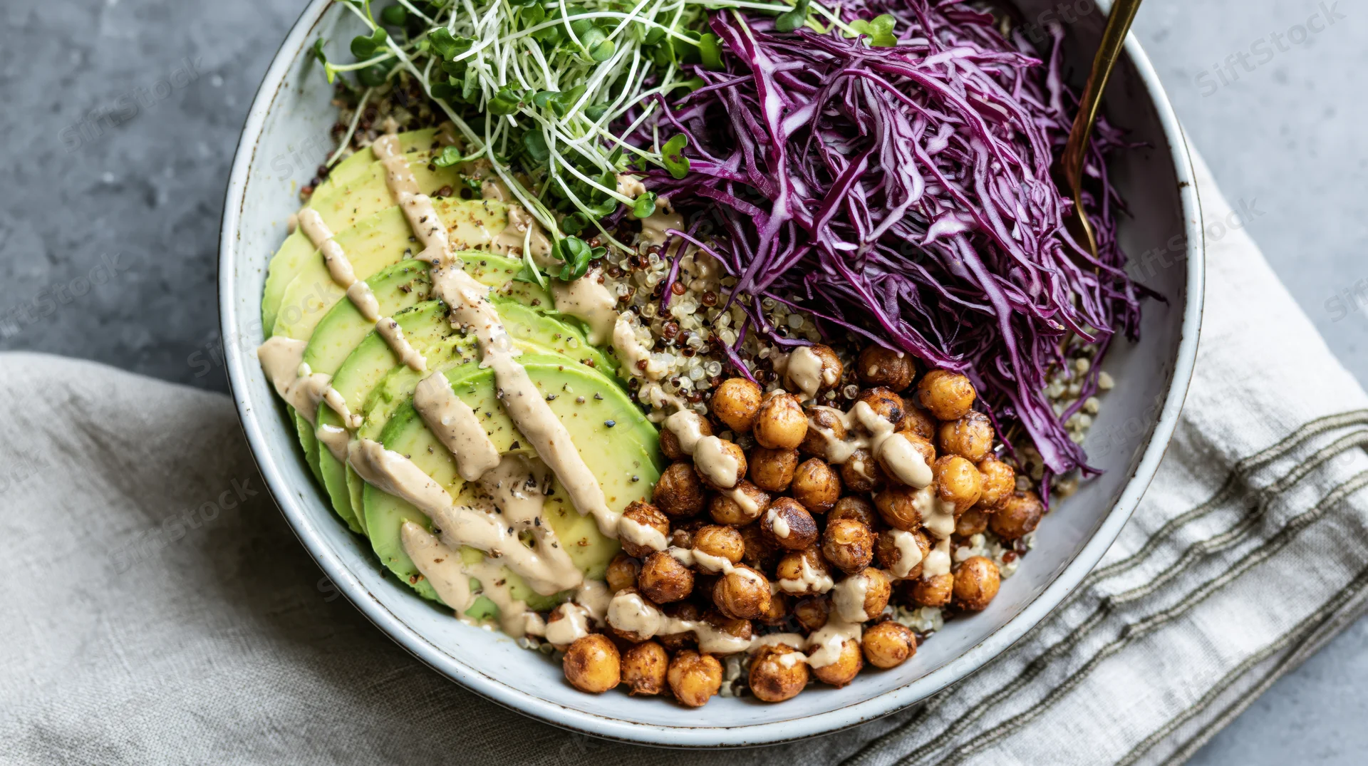 Overhead view of a grain bowl with quinoa, avocado, roasted chickpeas, purple cabbage, and sprouts in a ceramic dish on a gray surface with a linen napkin