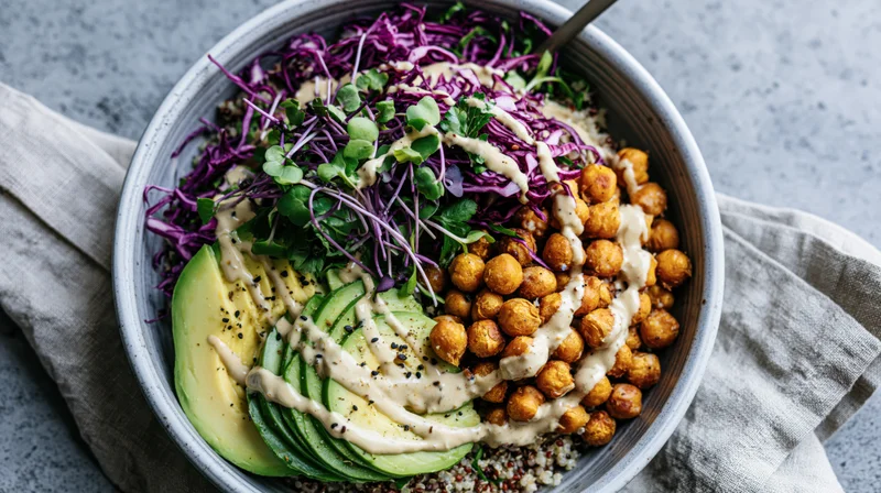 Overhead view of a grain bowl with avocado, chickpeas, purple cabbage and microgreens in a matte ceramic dish on a gray surface with a linen napkin