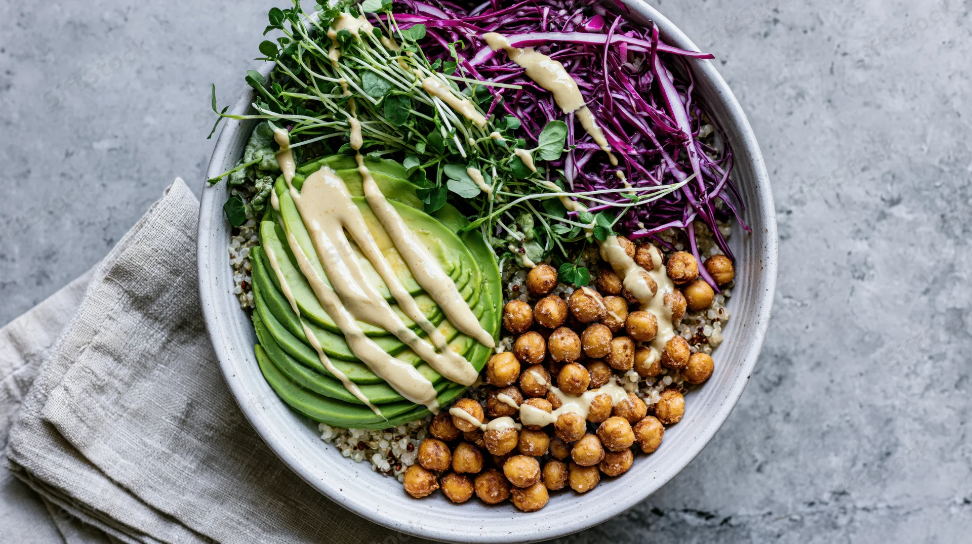 Overhead flat-lay of a grain bowl with avocado, chickpeas, quinoa, purple cabbage and microgreens in a matte ceramic dish on a gray stone surface with a linen napkin beside it