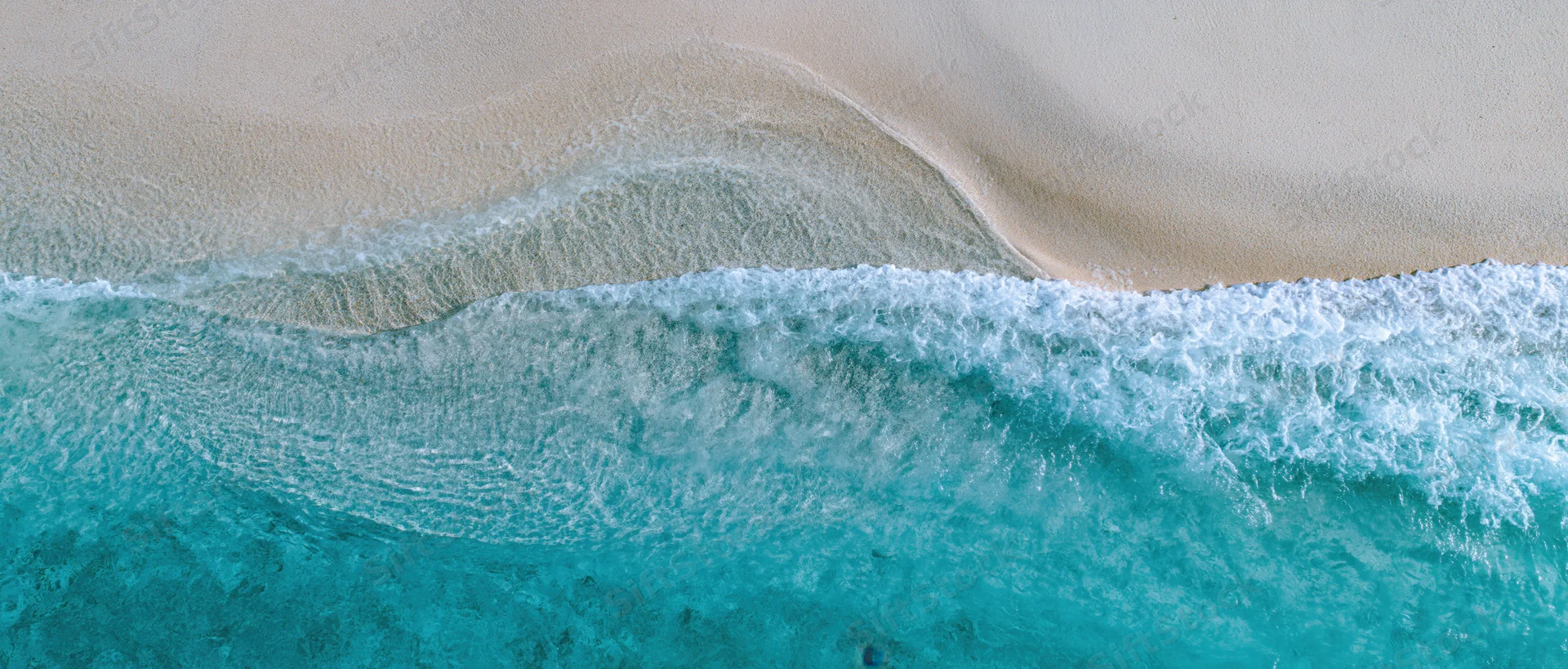 Overhead view of turquoise waves breaking on a pale sandy shoreline