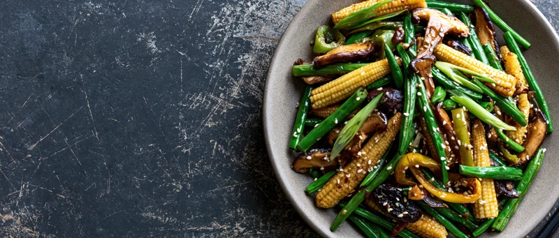 Stir-fried vegetables and mushrooms with baby corn and sesame seeds on a plate over dark textured surface