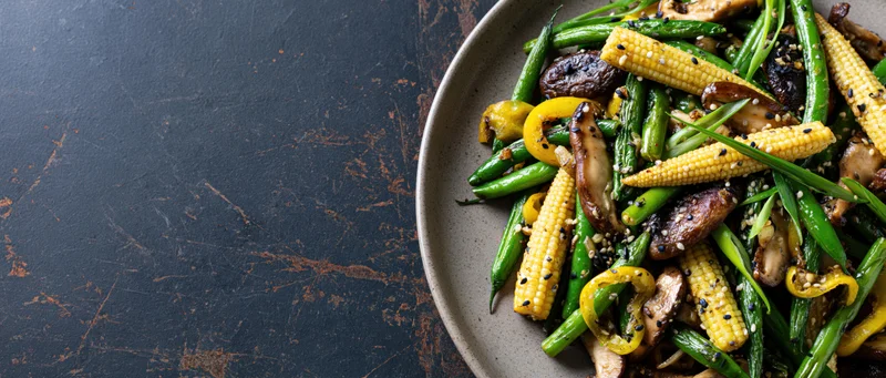 Stir-fried vegetables and mushrooms with corn and peppers on a plate on a dark textured surface