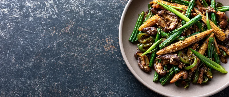 Stir-fried vegetables and mushrooms with baby corn and green onions on a plate over dark textured surface