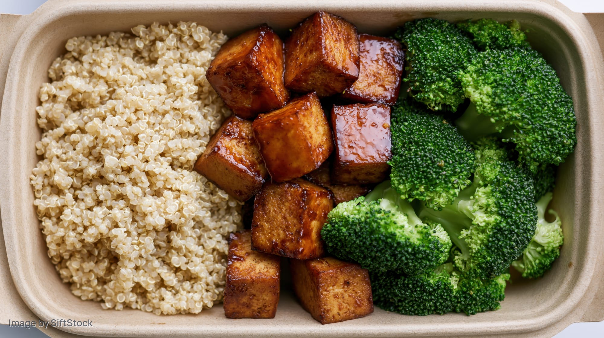 Overhead view of a divided kraft paper box with quinoa, glazed tofu cubes, and steamed broccoli arranged in sections
