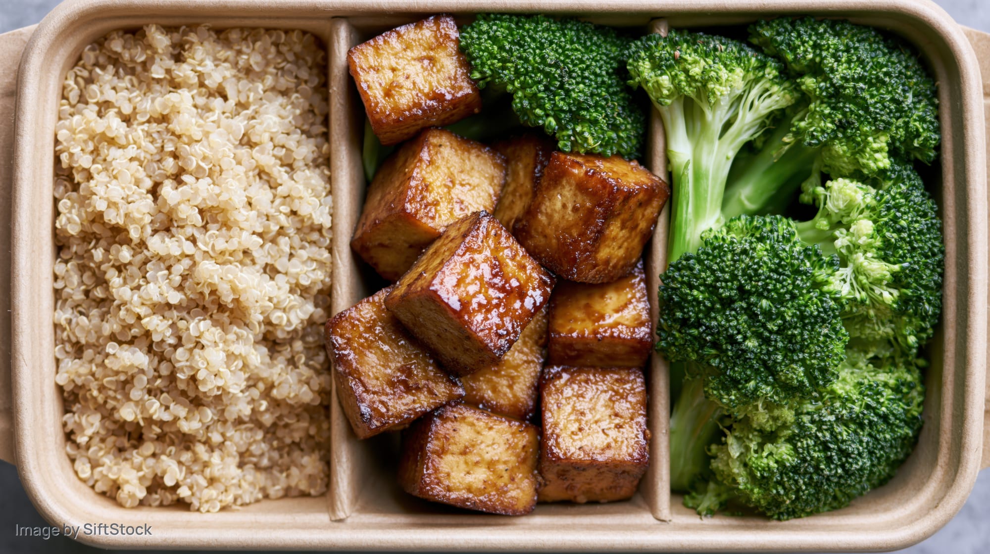 Overhead view of a divided kraft paper box containing quinoa, glazed tofu cubes, and steamed broccoli