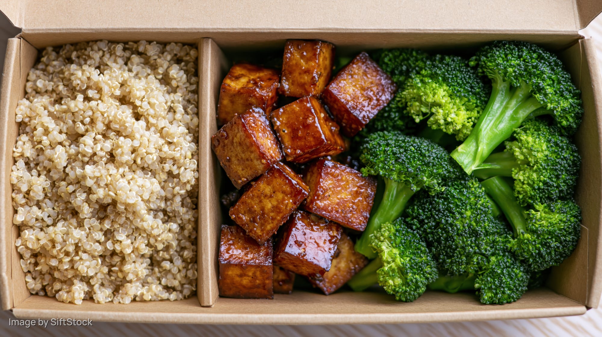 Overhead view of a divided kraft paper takeout box containing quinoa, glazed tofu cubes, and steamed broccoli