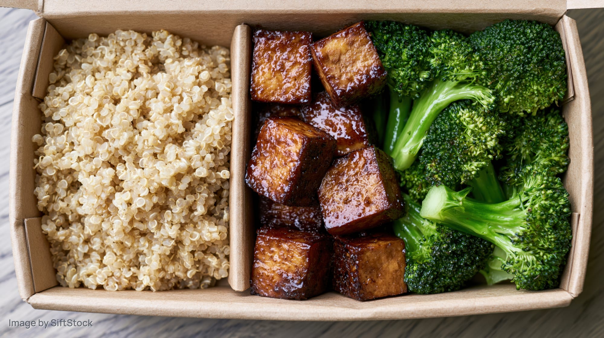 Overhead view of a divided kraft paper takeout box containing quinoa, glazed tofu cubes, and steamed broccoli