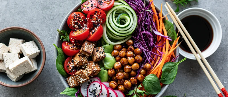 Overhead flat-lay of a vegan buddha bowl with tofu, chickpeas, zucchini, carrots, cabbage, tomatoes, spinach, radish, and sesame seeds on a gray stone surface with chopsticks and soy sauce on the side