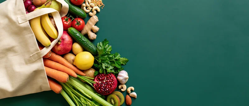 Cream canvas tote bag spilling fresh vegetables and fruits on green surface