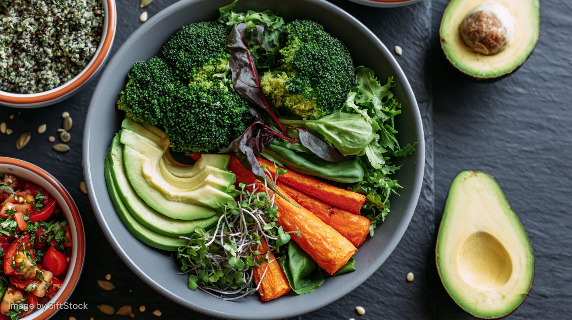 Overhead flat lay of a grey ceramic bowl filled with sliced avocado, broccoli, carrots, greens, and microgreens on a dark slate surface with side bowls of quinoa and tomato salad