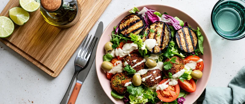 Overhead view of a pink bowl filled with falafel, grilled eggplant, and salad on a kitchen countertop