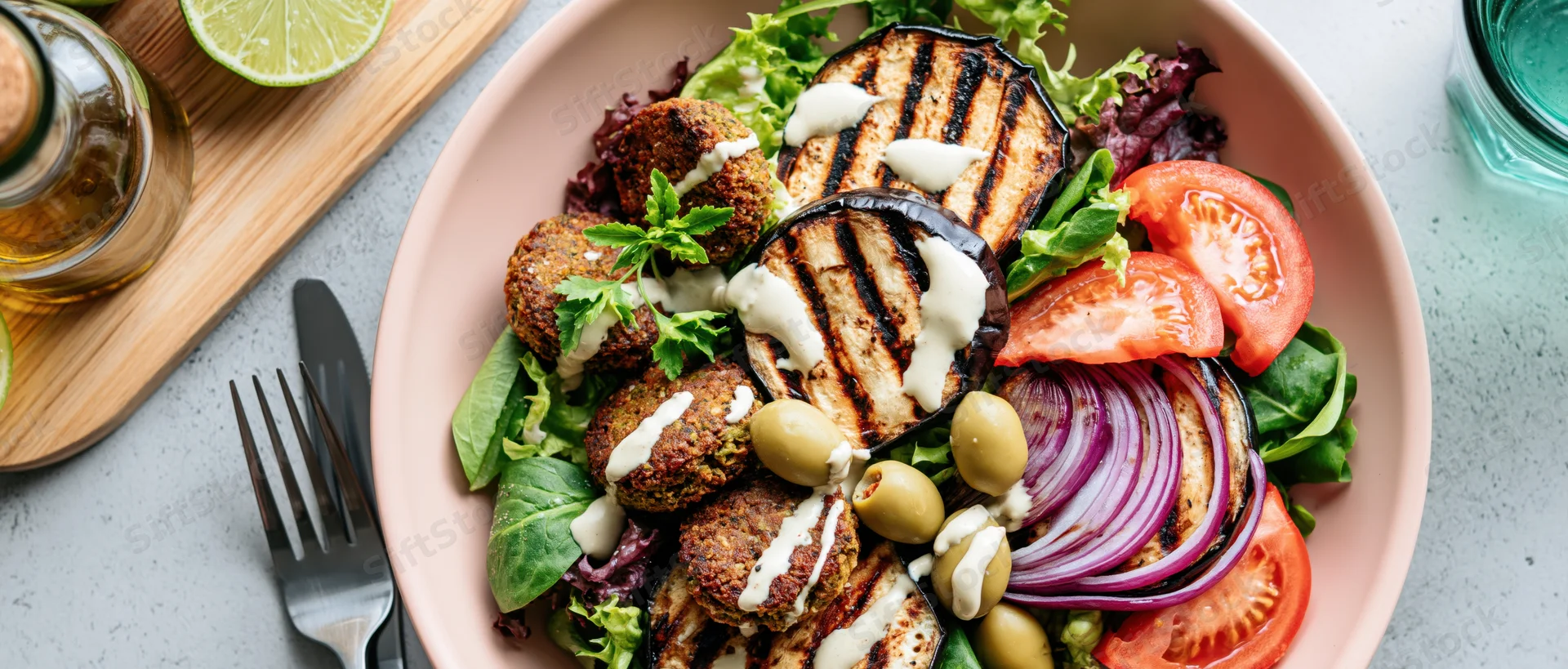 Overhead view of a pink bowl filled with falafel, grilled eggplant, tomatoes, onions, olives and parsley on a gray countertop